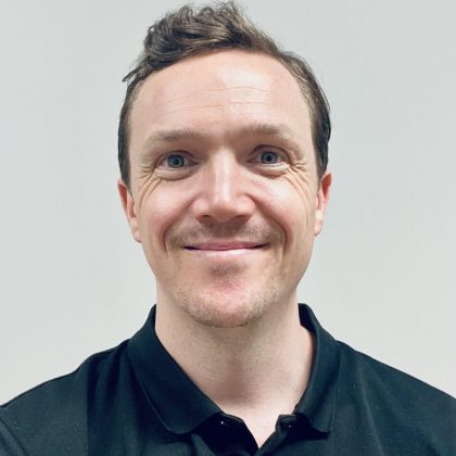 Lee Schneider smiles at the camera. He has short dark hair and in wearing a black polo shirt