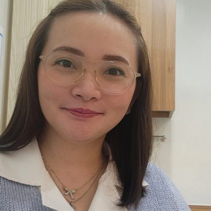 Dr Lucille Ocampo smiles at the camera. She has long dark hair and is wearing clear glasses and a white and blue shirt