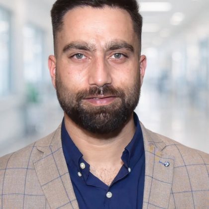 Dr Gagandeep singh smiles at the camera. he has short dark hair and is wearing is navy shirt with a beige checkered blazer
